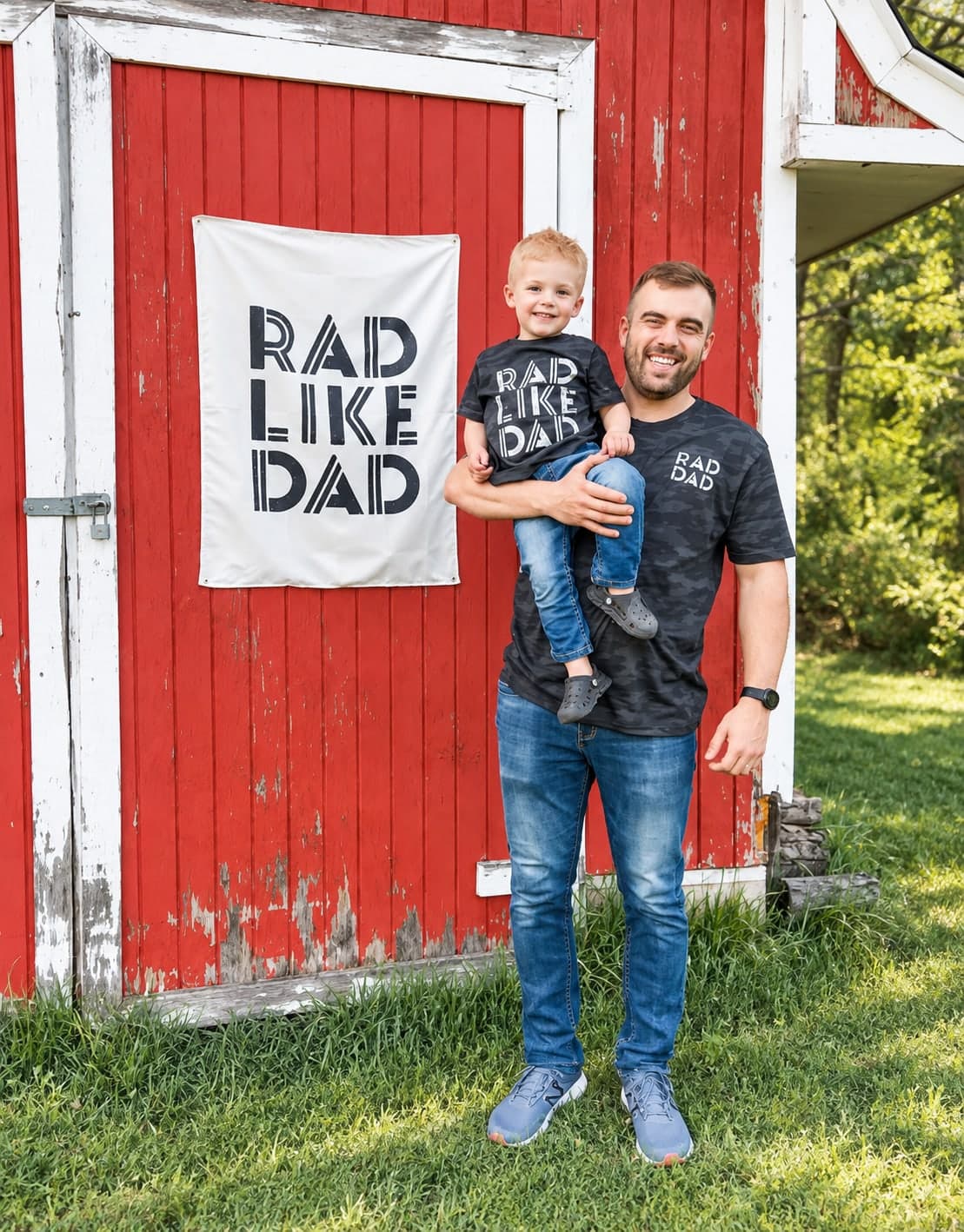 Founder holding his son, both in matching Rad Like Dad tees, in front of a red barn